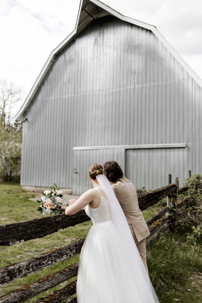 Bride and groom lean on a fence and look at the blue barn at this Estate 248 wedding in Langley, British Columbia