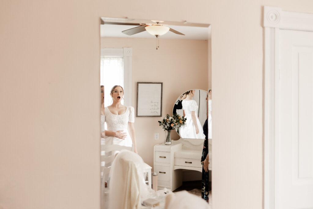 The bride reacts with an open mouth of shock looking at herself in the mirror in her wedding dress at this Estate 248 wedding in Langley, British Columbia