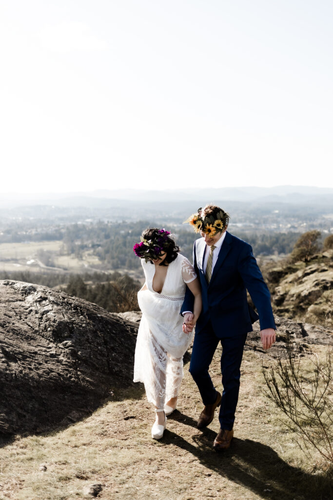 The couple hold hands and smile as they walk together in front of a view of the city at their Mount Douglas elopement.