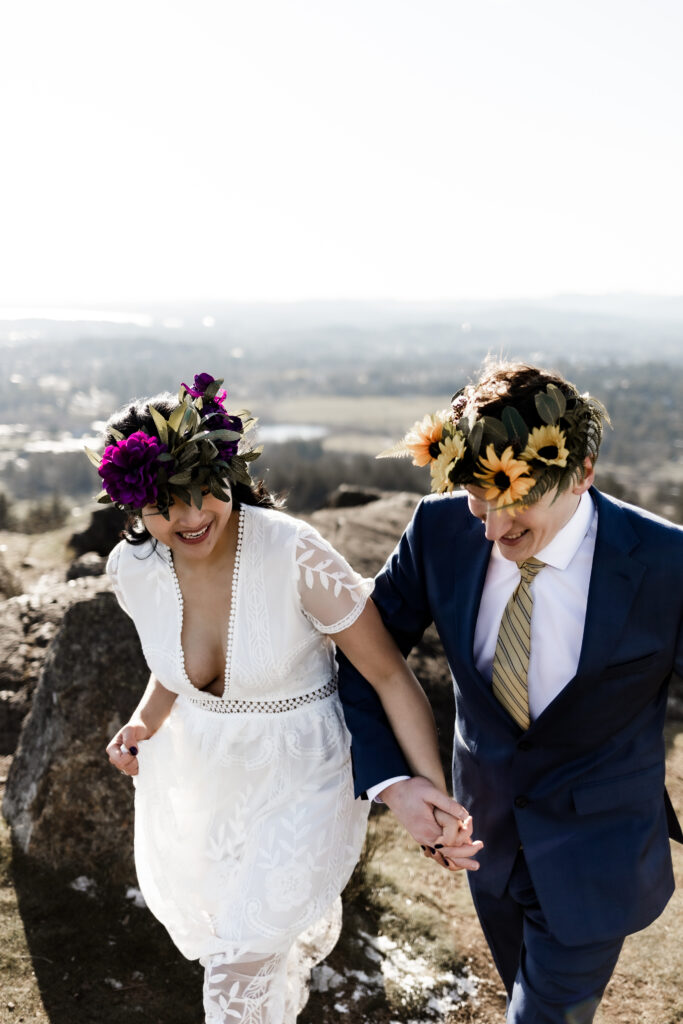 The couple hold hands and smile as they walk together in front of a view of the city at their Mount Douglas elopement.