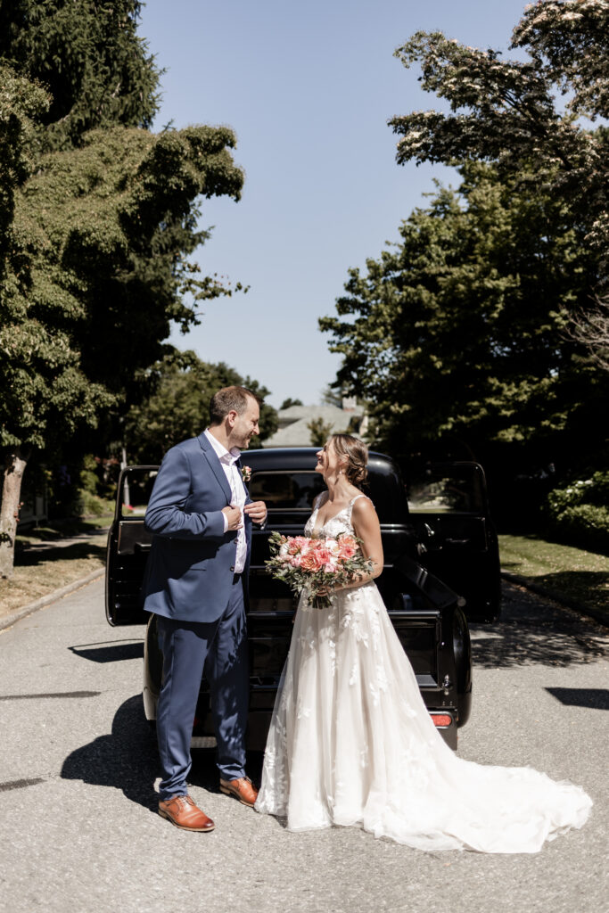 the bride and groom smile at each other in front of the black vintage truck at their garden party wedding