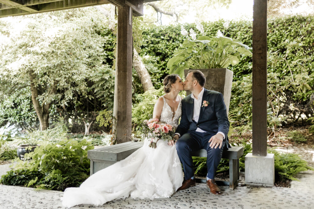 The newly couple shares a kiss, sitting beside each other on a bench at their garden party wedding