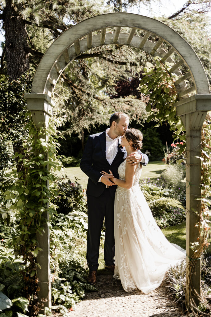 The groom gives the bride a kiss on her forehead as they stand beneath an archway at their garden party wedding