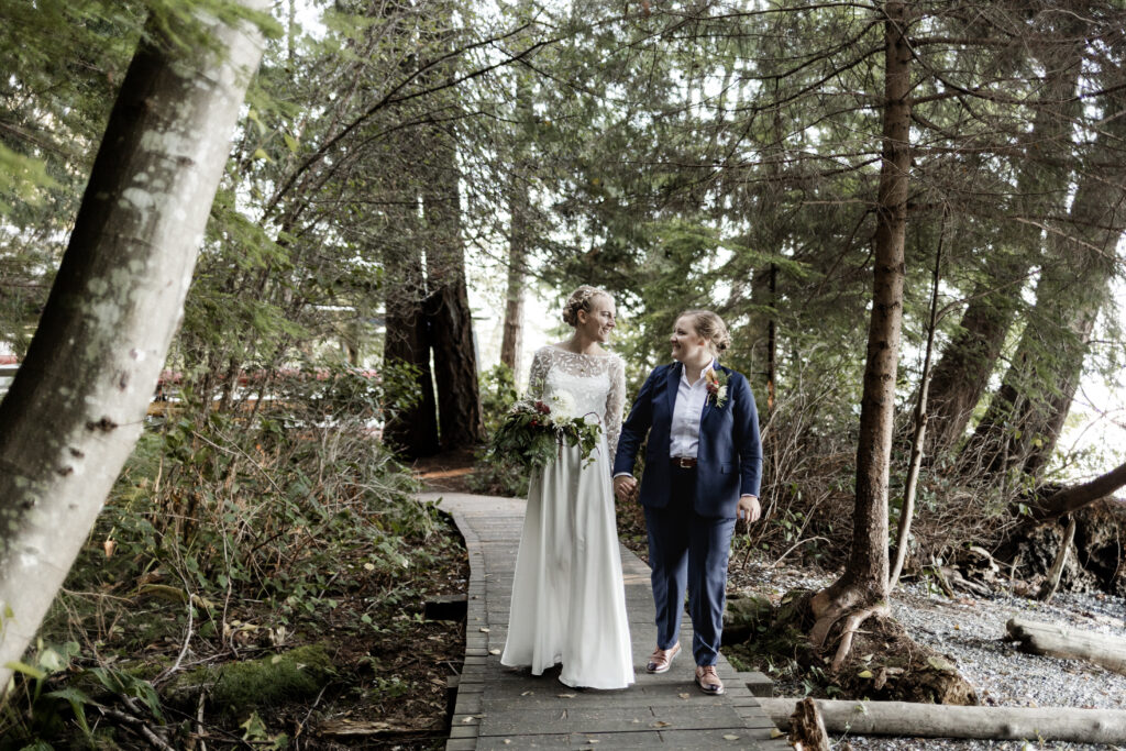 The couple walk hand in hand down a wooden boardwalk at this Camp Elphinstone wedding.