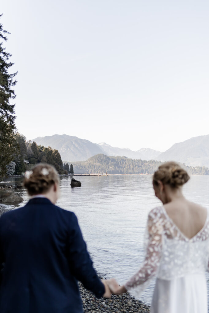 The couple hold hands, framing the mountains behind them and showing off the lake at this Camp Elphinstone wedding.