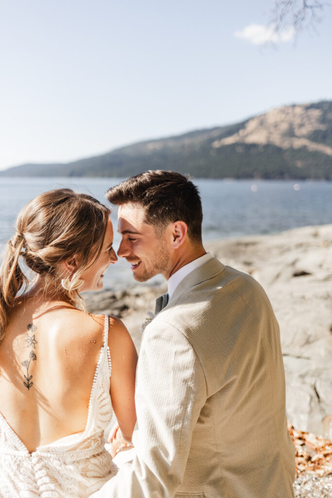 The newly weds touch noses as they sit infront of an ocean and mountain view on the beach at this Kildara Farms wedding