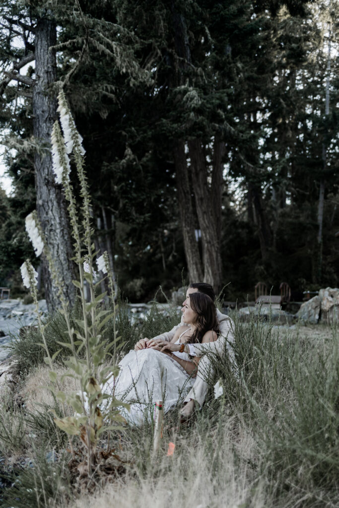 The couple share a cuddle sitting amongst the tall green grass on the edge of the beach at their east sooke elopement.