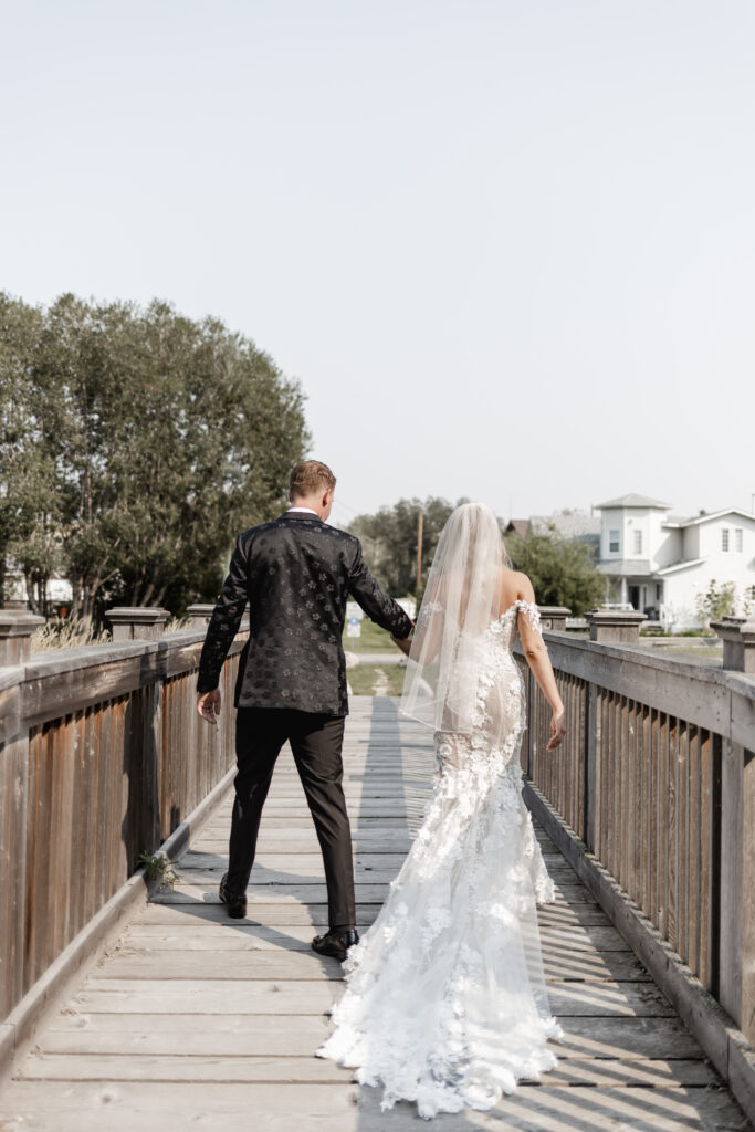 Bride and groom walk holding hands on a wooden bridge at this lakeside wedding at Alberta Beach