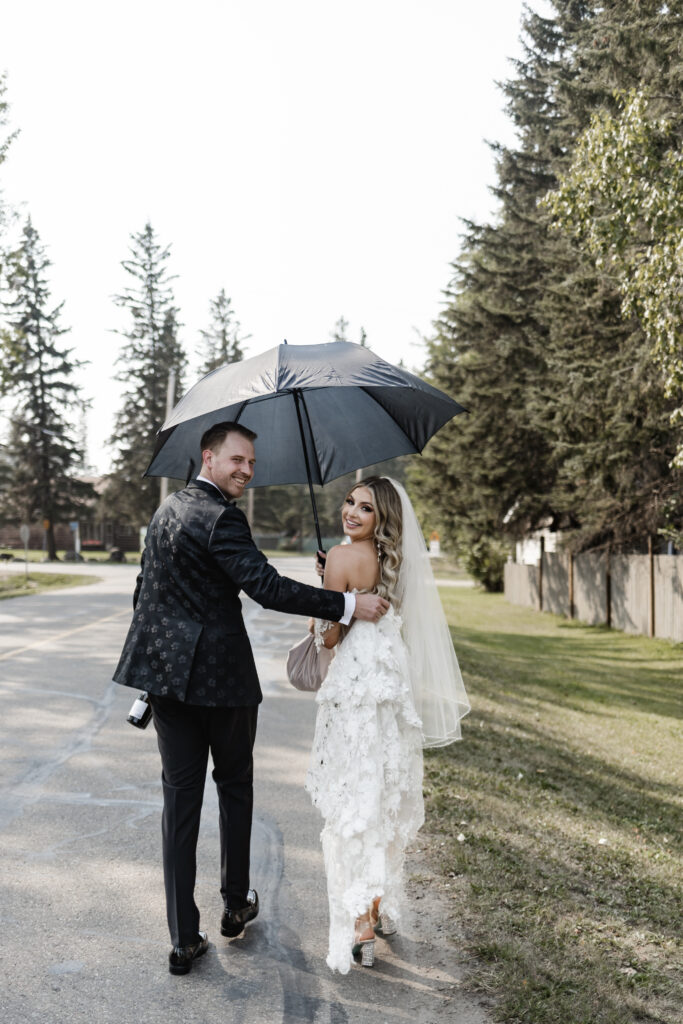 bride and groom look back as they walk underneath an umbrella at this lakeside wedding at Alberta Beach