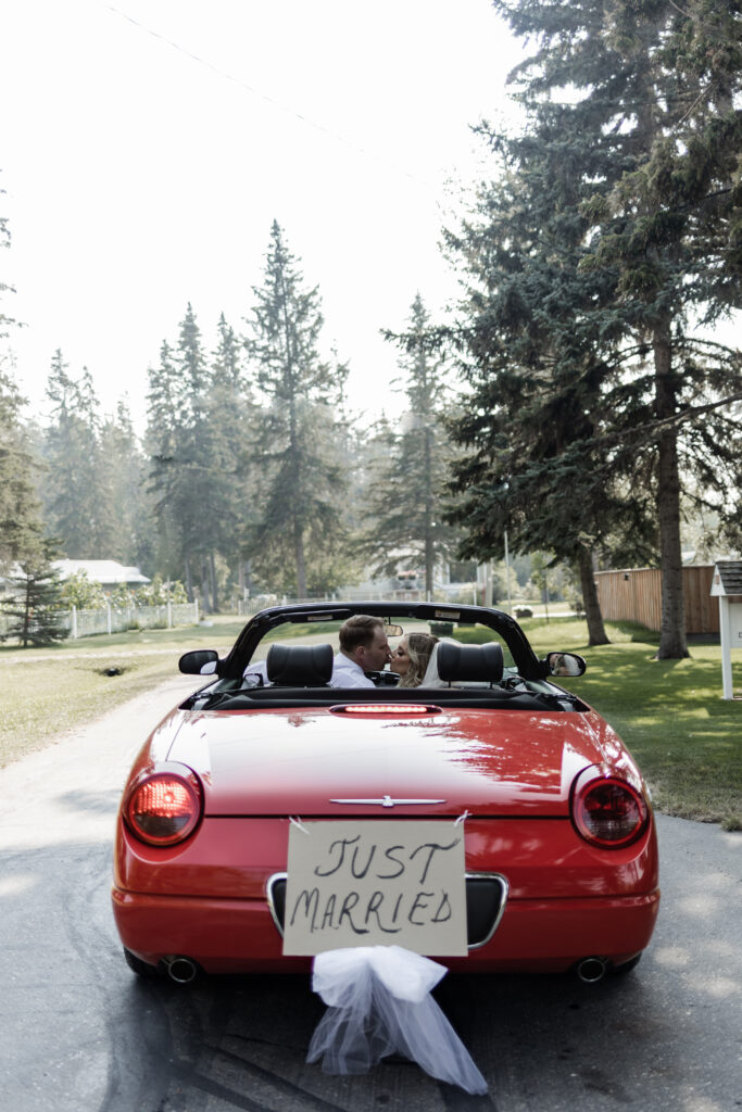 bride and groom share a kiss as they sit in a red car with a just married sign on the back at this lakeside wedding at Alberta Beach