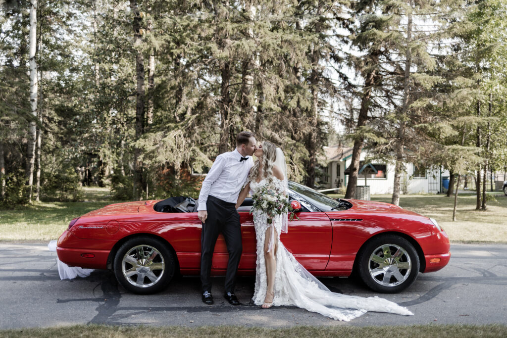 bride and groom share a kiss as they stand in front of the red car at this lakeside wedding at Alberta Beach