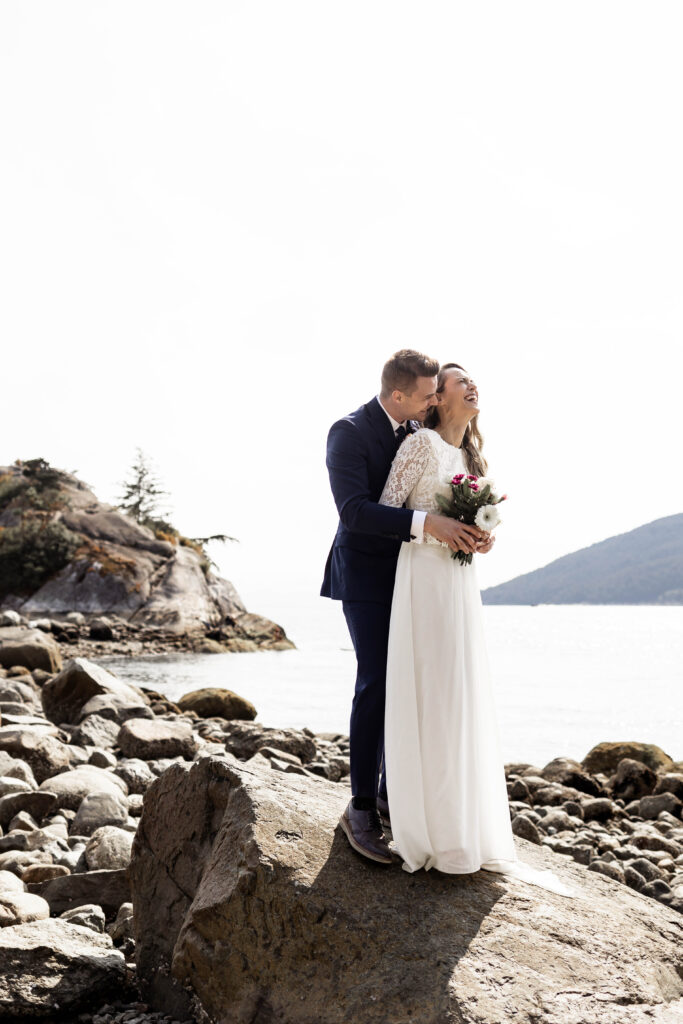 Groom hugs bride from behind while they stand on a rock at this Whytecliff Park elopement