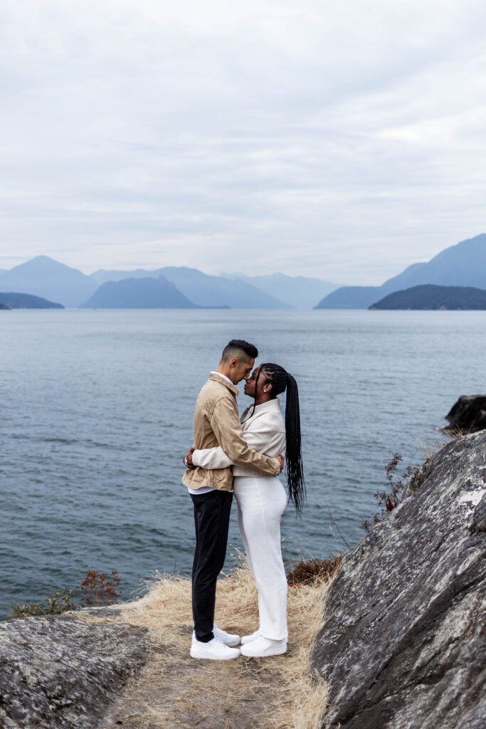 The couple stand nose to nose in front of blue ocean and mountain views at this Whytecliff Park engagement shoot