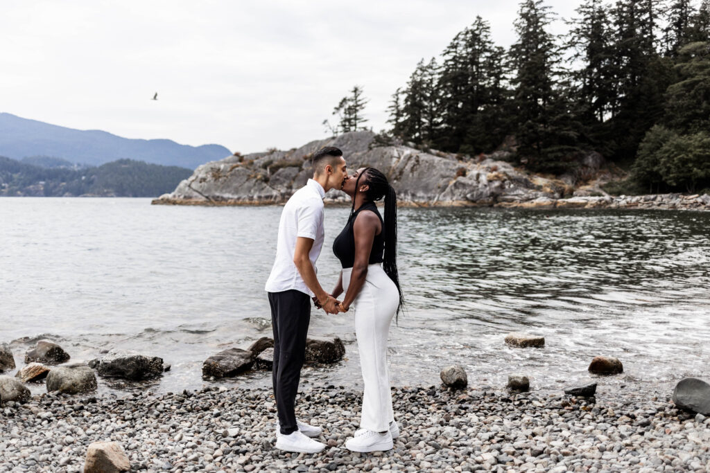 A couple hold hands and kiss in front of the water at this Whytecliff Park engagement shoot