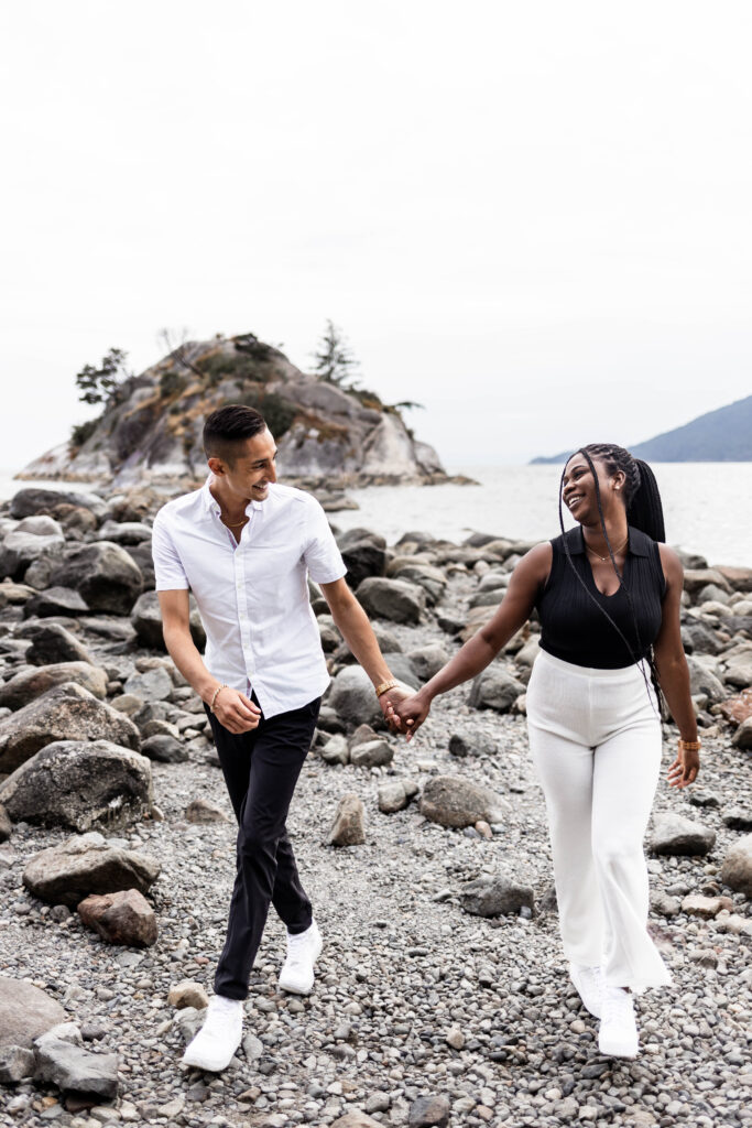 The couple hold hands as they smile at each other and walk on the beach at this Whytecliff Park engagement shoot