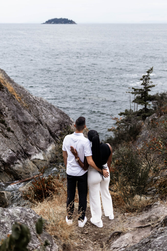 The couple stand, arms around each other, looking at the ocean view at this Whytecliff Park engagement shoot