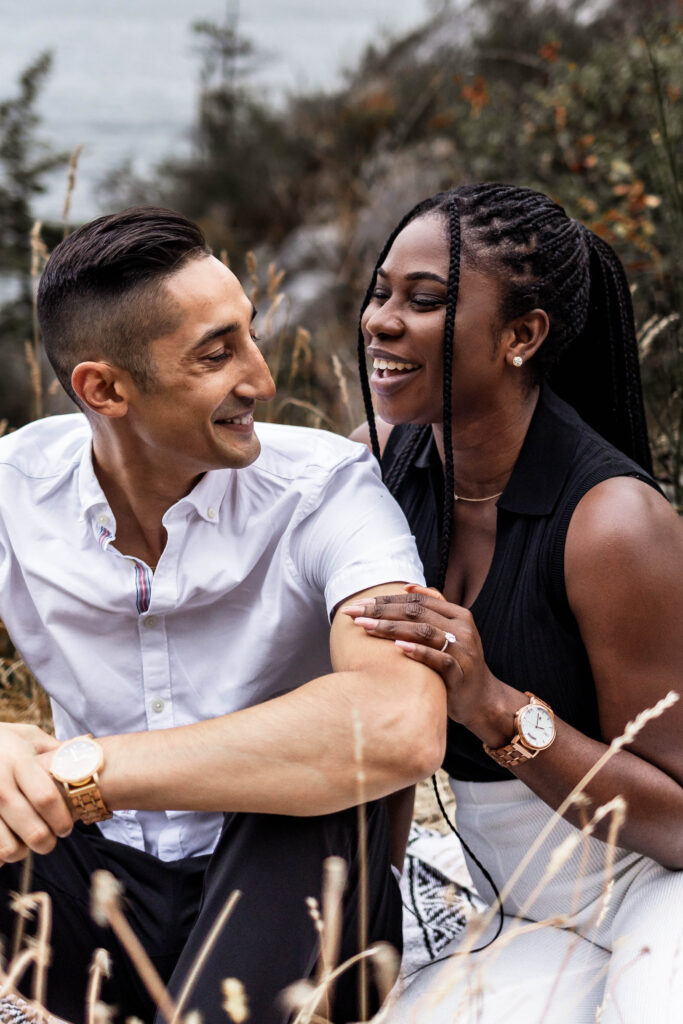 The couple sit beside each other and share a laugh at this Whytecliff Park engagement shoot