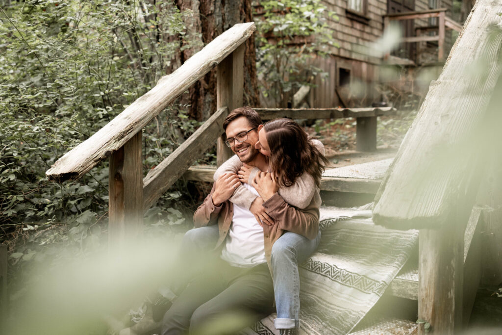 Stef hugs Ben from behind in the forest at this Lighthouse Park Engagement Shoot