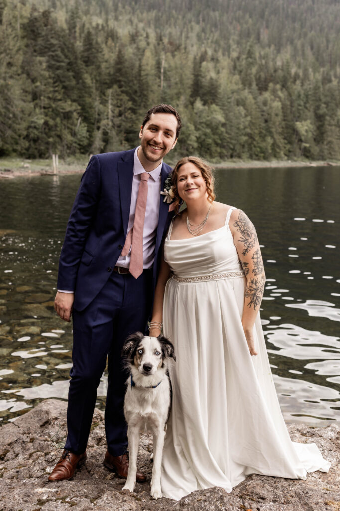 Bride, groom, and dog pose for a family photo in front Mabel Lake at their elopement