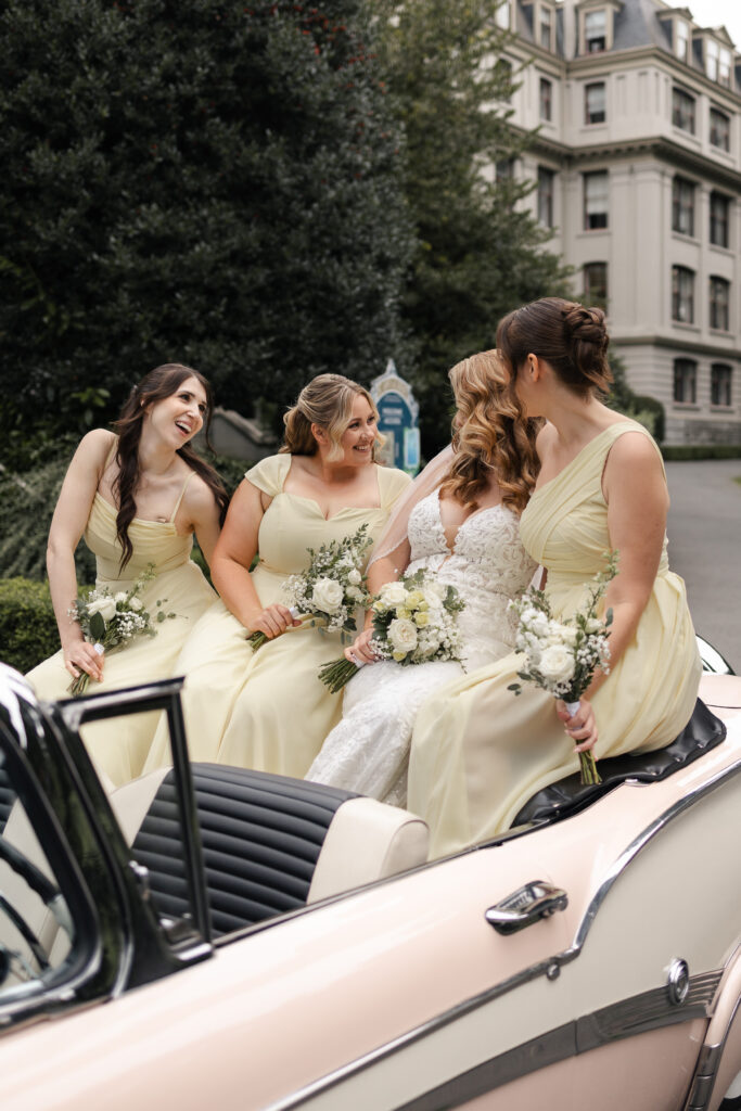 Bride and bridesmaids giggle at each other while posed on the back of a pink vintage car at this Victoria cathedral wedding